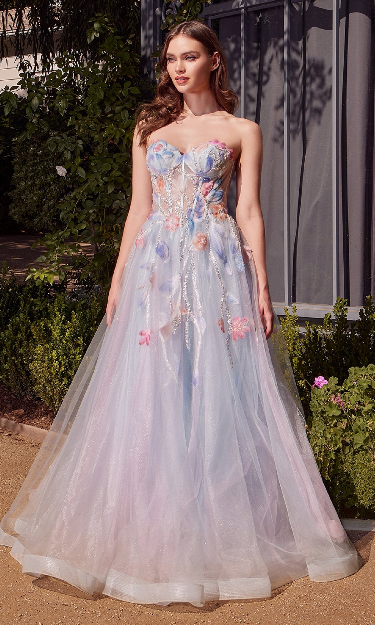 A woman stands outdoors on a dirt path near greenery and a building, wearing the Andrea & Leo A1383 long strapless prom dress with glitter and floral embellishments.